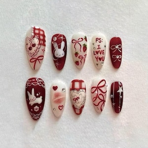May include: A set of ten artificial nails with a variety of designs. The nails feature a mix of white and burgundy colors, with illustrations of hearts, bows, strawberries, and a bunny. Some nails have the text "PS: I LOVE YOU".