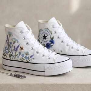 May include: A pair of white high-top platform sneakers featuring embroidered blue and purple wildflowers, green stems, and a small bee on the side. The shoes include white laces, a black stripe on the sole, and a circular Converse All Star logo patch.