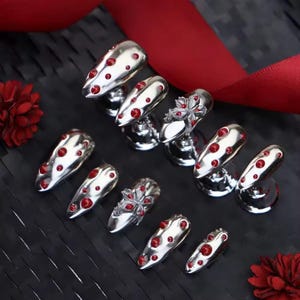 Peut inclure: Ongles décoratifs argentés ornés de pierres précieuses rouges. Les designs d'ongles sont présentés sur de petits supports argentés, dont certains avec un motif floral. Un ruban rouge et une surface texturée sombre composent l'arrière-plan.