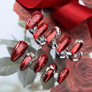 May include: A selection of red, glitter-infused artificial nails, some with silver decorations, presented on small chrome stands. The nails are set against a floral background and a red ribbon.