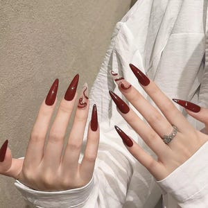 May include: Long, pointed, burgundy-coloured artificial nails. One nail features a clear design with a swirling pattern in a darker shade of burgundy. A silver ring with a floral design is visible.