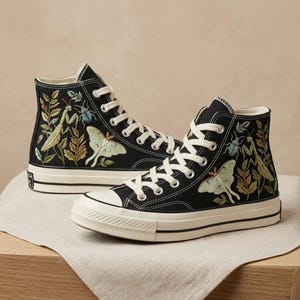 Könnte beinhalten: Ein Paar schwarze High-Top-Sneaker aus Canvas mit aufwendigen Stickereien von grünen Gottesanbeterinnen, blassgrünen Mondmotten, blauen Käfern und verschiedenen Pflanzenmotiven. Die Schuhe haben weiße Schnürsenkel, weiße Gummikappen und weiße Sohlen.
