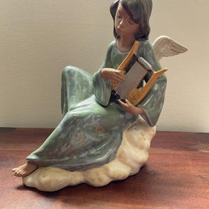 May include: A porcelain figurine of an angel with dark hair, wearing a flowing sage green robe with a glossy finish. The angel is seated on a cream-colored cloud, playing a small tan-colored harp with visible strings, and has white wings.