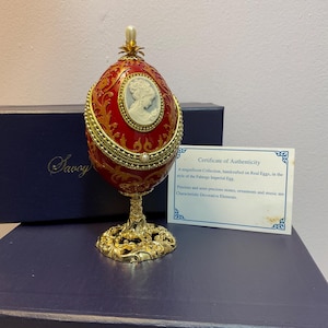 Op de afbeelding: Een rood en goud decoratief ei in de stijl van een Fabergé Imperial Egg, met een witte cameo en parelaccenten. Het ei staat op een gouden filigraanstandaard, met een certificaat van echtheid.
