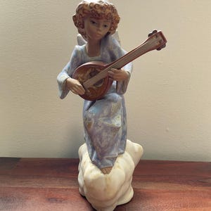 May include: A porcelain figurine of an angel with curly brown hair, wearing a light blue and lavender patterned robe, standing on a white cloud base while playing a brown wooden lute. The angel has visible wings and a serene facial expression.