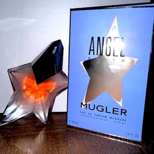 May include: A star-shaped perfume bottle with a black cap and orange liquid inside, next to a blue box with the word "ANGEL" and a star design. The box also says "MUGLER" and "1.6 Fl. Oz."