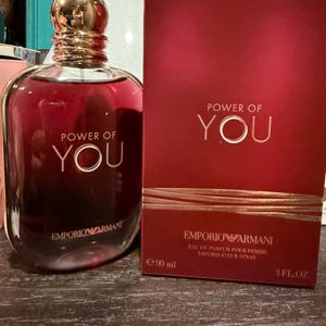May include: A red-tinted glass perfume bottle with a gold cap and a matching red box. The bottle and box both feature the text "POWER OF YOU" and the Emporio Armani logo. The bottle contains 3 fl oz of perfume.