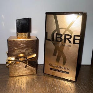 May include: A gold-toned perfume bottle with a black cap and a matching box. The box features the word "LIBRE" in large black letters and the brand name "Yves Saint Laurent". The perfume bottle has a gold bow.