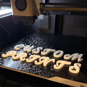 May include: A 3D printer creating white plastic letters spelling "CUSTOM PRINTS" on a black patterned surface. The printer head is visible above the letters, suggesting a custom printing service.