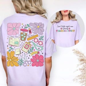 May include: A lilac t-shirt with a colourful graphic on the back featuring flowers, school supplies, and the text "Lesson Plans". A smaller image shows the front of the shirt with the text "Can't talk right now Doing Teacher Stuff".