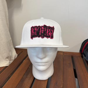 Può includere: Un cappellino da baseball bianco con una grafica sul pannello frontale che riporta la parola MONEY in lettere nere e rosa in grassetto. Il design incorpora un'immagine dettagliata di una banconota da cento dollari all'interno delle lettere.