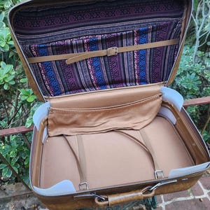 May include: Open vintage suitcase with a brown exterior and a patterned interior in shades of purple, blue, and tan. The suitcase has a leather handle and straps, and a metal zipper. The interior features a pocket and straps to secure items.
