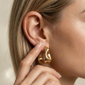 Könnte beinhalten: Nahaufnahme eines Ohrs mit einem Paar goldfarbener, unregelmäßiger Creolen. Die Ohrringe haben eine strukturierte Oberfläche und eine polierte Oberfläche. Die Person hat helle Haut und blondes Haar.