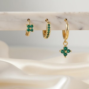 Puede incluir: Tres pendientes de aro dorados con piedras preciosas verdes. Uno presenta un diseño de trébol de cuatro hojas, otro tiene gemas verdes alrededor del aro, y el tercero tiene un dije de trébol. Los pendientes se exhiben sobre una superficie de mármol blanco.