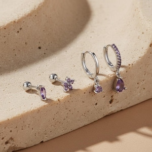 Könnte beinhalten: Eine Auswahl an silbernen Ohrringen mit lila Akzenten. Das Set umfasst einen Ohrstecker mit einem lilafarbenen Stein im Marquiseschliff, einen blütenförmigen Ohrstecker, kleine Creolen mit einem hängenden lila Stein und eine Creole mit einer Reihe lila Steine und einem tropfenförmigen Stein.