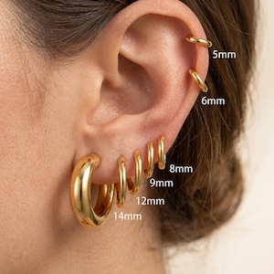 Puede incluir: Primer plano de una oreja adornada con una colección de pendientes de aro dorados. Los pendientes varían en tamaño, de 5 mm a 14 mm, mostrando un estilo graduado. El acabado dorado pulido refleja la luz, resaltando el diseño liso y redondeado.