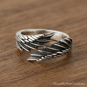 Puede incluir: Un anillo de plata con un dise&ntilde;o abierto con detalles de alas. El anillo est&aacute; sobre una superficie de madera, mostrando los intrincados detalles de las plumas. Es una pieza de joyer&iacute;a.