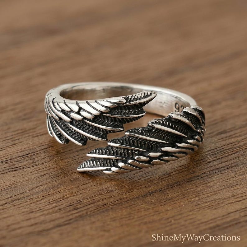 Puede incluir: Un anillo de plata con un dise&ntilde;o abierto con detalles de alas. El anillo est&aacute; sobre una superficie de madera, mostrando los intrincados detalles de las plumas. Es una pieza de joyer&iacute;a.