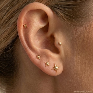 Pode incluir: Um conjunto de cinco brincos de pino dourados em forma de estrela dispostos ao longo do lóbulo e da cartilagem. Cada brinco apresenta um design de estrela de várias pontas com um ponto central. O texto ShineMyWayCreations é visível.