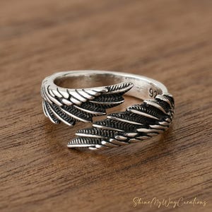 Puede incluir: Un anillo plateado con un dise&ntilde;o abierto, con detalles en forma de alas. El anillo tiene un acabado pulido y est&aacute; sobre una superficie de madera. El n&uacute;mero "92" es visible en el anillo.