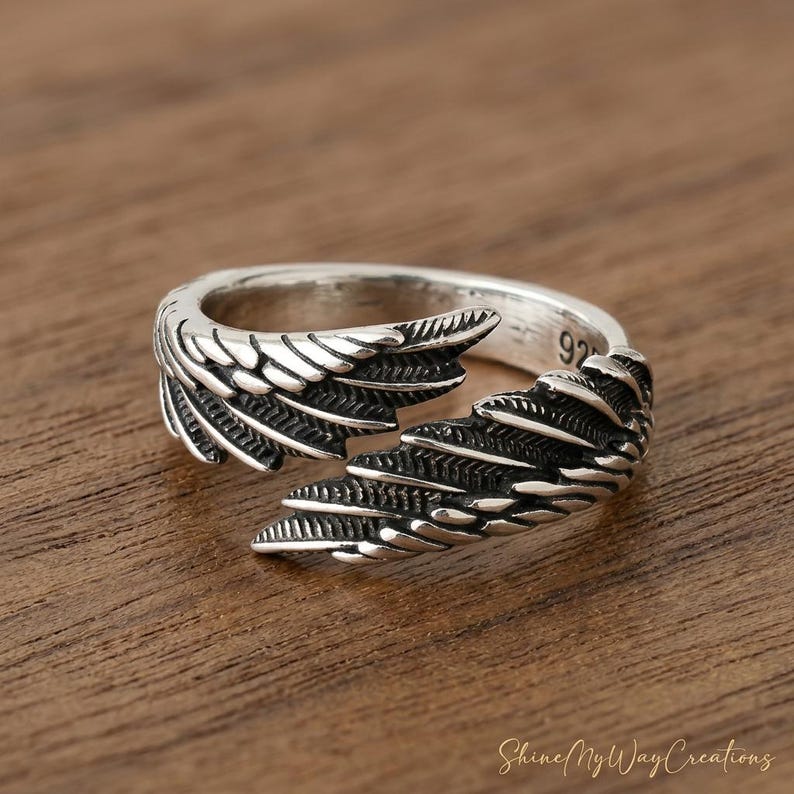 Puede incluir: Un anillo plateado con un dise&ntilde;o abierto, con detalles en forma de alas. El anillo tiene un acabado pulido y est&aacute; sobre una superficie de madera. El n&uacute;mero "92" es visible en el anillo.