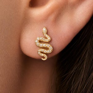 Könnte beinhalten: Ein goldfarbener Ohrstecker in Form einer gewundenen Schlange. Die Oberfläche der Schlange ist mit zahlreichen kleinen, klaren, funkelnden Steinen besetzt, die dem geschwungenen, S-förmigen Körper vom Kopf bis zum Schwanz folgen.