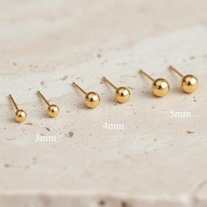 Puede incluir: Un conjunto de pendientes de botón dorados con forma de bola, en varios tamaños: 3 mm, 4 mm y 5 mm. Los pendientes se muestran sobre una superficie clara, resaltando su acabado pulido y brillante. La imagen destaca los diferentes tamaños disponibles.
