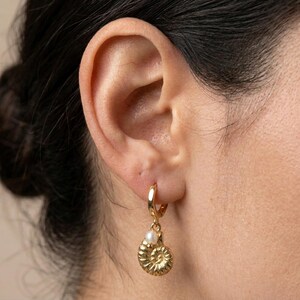 Peut inclure: Une boucle d'oreille créole dorée avec une breloque pendante en forme de fossile d'ammonite texturé. Une petite perle blanche ronde est fixée au sommet de la breloque dorée, reposant juste en dessous de l'anneau circulaire fixé au lobe.