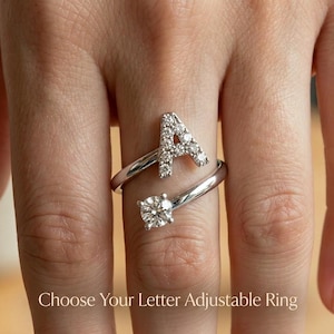 Puede incluir: Anillo ajustable plateado con la letra "A" adornada con pequeñas piedras transparentes. Una piedra grande, redonda y transparente está engastada en el extremo opuesto. El texto "Choose Your Letter Adjustable Ring" es visible en la parte inferior.