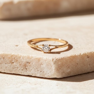 Puede incluir: Un anillo de oro con una sola gema redonda y transparente. El anillo está colocado sobre una superficie texturizada de color claro. La banda tiene un diseño abierto a ambos lados de la gema.