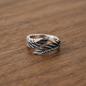 Puede incluir: Un anillo plateado con un dise&ntilde;o abierto, con intrincados patrones de alas en cada lado. El anillo se encuentra sobre una superficie de madera c&aacute;lida, mostrando su intrincada artesan&iacute;a. El anillo es talla 92.