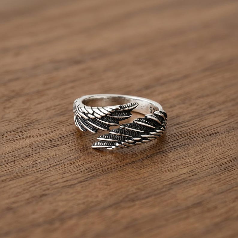 Puede incluir: Un anillo plateado con un dise&ntilde;o abierto, con intrincados patrones de alas en cada lado. El anillo se encuentra sobre una superficie de madera c&aacute;lida, mostrando su intrincada artesan&iacute;a. El anillo es talla 92.