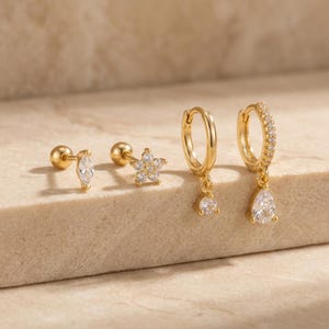 Könnte beinhalten: Eine Sammlung goldfarbener Ohrringe. Das Set beinhaltet einen Ohrstecker im Marquise-Schliff, einen blütenförmigen Ohrstecker und zwei Creolen mit tropfenförmigen Anhängern. Ein Reifen ist mit kleinen Edelsteinen besetzt.