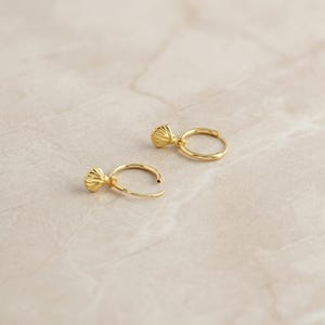 Puede incluir: Un par de pendientes de aro dorados con un dije texturizado en forma de concha. Los pendientes se muestran sobre una superficie clara, similar al mármol. Los pendientes están abiertos, mostrando el mecanismo de cierre.