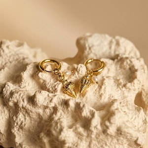 May include: A pair of gold hoop earrings featuring a dangling textured seashell charm and a small white pearl bead. The earrings are crafted with a polished gold finish and a circular hoop design, showcasing intricate shell details and a lustrous pearl.
