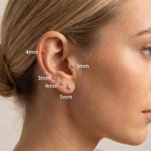 Könnte beinhalten: Nahaufnahme eines Ohrs mit mehreren goldenen Stern-Ohrsteckern. Die Ohrringe sind in den Größen 3 mm, 4 mm und 5 mm erhältlich. Das Ohr ist vor einem verschwommenen Hintergrund platziert.
