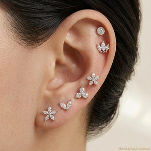 Puede incluir: Primer plano de una oreja adornada con múltiples pendientes de botón. Los pendientes presentan varios diseños, incluyendo formas de flores, una corona y un grupo de pequeñas piedras. Los pendientes son de color plateado y parecen estar hechos de metal y piedras preciosas.