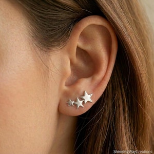 Puede incluir: Un pendiente trepador plateado con tres estrellas de cinco puntas de diferentes tamaños dispuestas en fila. Las estrellas tienen un acabado liso y pulido, siguiendo la curva natural del lóbulo de la oreja. El texto ShineMyWayCreations es visible.