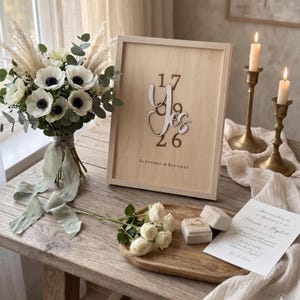 Peut inclure: Un panneau en bois encadré avec le mot "Yes" et les chiffres "17" et "26" dans une police décorative. Un bouquet de fleurs blanches avec des accents verts et un ruban, ainsi qu'un petit bouquet de roses blanches, sont sur une table en bois.
