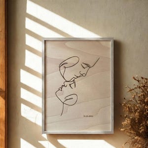 Peut inclure: Une œuvre d'art encadrée représentant un dessin au trait minimaliste de deux visages enlacés. L'œuvre est à l'encre noire sur un fond clair en grain de bois, avec la date 12.10.2021 imprimée en bas. Le cadre est argenté.