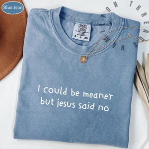Könnte beinhalten: Ein blaues Jeans-T-Shirt mit dem Text "I could be meaner but jesus said no" in Weiß. Das Shirt hat einen Rundhalsausschnitt und kurze Ärmel. Eine goldene Halskette mit Anhänger liegt über dem Shirt.