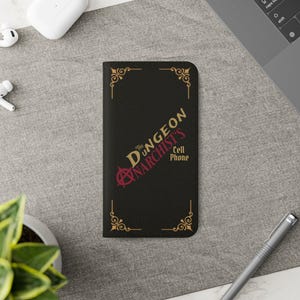May include: A black rectangular phone case featuring gold decorative corner flourishes and the text "THE DUNGEON NARCHIST'S CELL PHONE" in gold and red lettering, with a red anarchy symbol integrated into the first letter of the word NARCHIST'S.