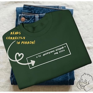 May include: Dark green t-shirt with a white rectangular graphic containing mirrored text. The shirt is folded on top of a pair of blue jeans. Text on the image reads "READS CORRECTLY IN MIRROR!" with an arrow pointing to the graphic.