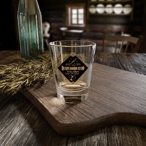 May include: A clear shot glass with the words "Desperado Club" in gold lettering on a black diamond-shaped label. The glass sits on a wooden cutting board with a bottle and wheat stalks in the background.