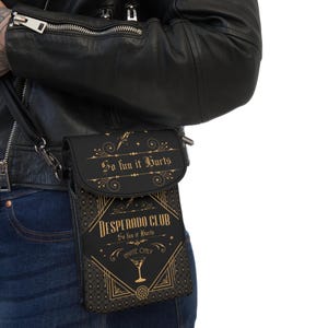Könnte beinhalten: Schwarze Umhängetasche mit goldenem Text "So fun it Hurts" und "Desperado Club". Die Tasche hat einen Klappverschluss und ein dekoratives Design mit einem Cocktailglas-Symbol. Die Tasche wird über der Schulter getragen.