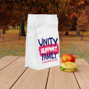 May include: A white lunch bag featuring a dark blue graphic design with the text UNITY SUPPORT FAMILY & KNEECAPPING B*TCHES printed over a pink brushstroke background. The bag is positioned next to a red apple and a sandwich on a wooden surface.