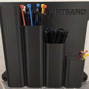 Puede incluir: Organizador de bridas de plástico negro con la palabra "BUNTBAND" en relieve en la parte posterior. El organizador tiene cuatro compartimentos llenos de bridas negras, rojas, azules y amarillas. Diseñado para sujetar y organizar bridas.