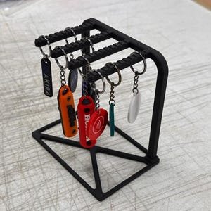 Pode incluir: Um porta-chaves de metal preto com vários chaveiros pendurados. Os chaveiros são de várias cores e formas, incluindo laranja, vermelho e branco. Um chaveiro tem a palavra "AKIRA".