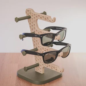 May include: A beige and olive green sunglasses display stand holding two pairs of black sunglasses. The stand has a honeycomb design and is made of plastic. The sunglasses have dark frames and green lenses.
