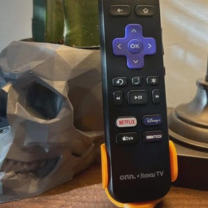 Puede incluir: Un mando a distancia de TV Roku negro con botones direccionales morados y botones de servicio de transmisión, en un soporte naranja. A la izquierda, una maceta gris con forma de calavera con una planta verde.